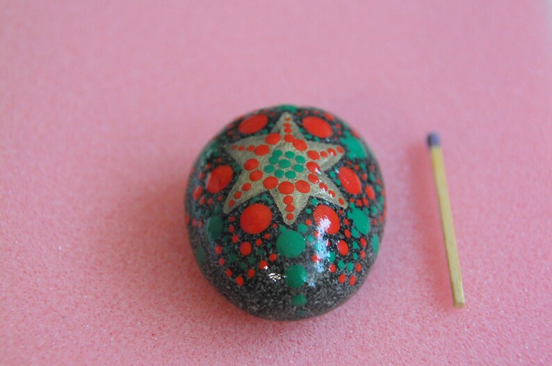 May include: A hand-painted rock with a green and red star design. The rock is about the size of a matchstick.