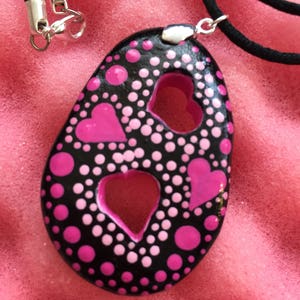 May include: Black pendant necklace with a heart-shaped cutout and pink polka dots and hearts. The pendant is on a black cord with a silver clasp. The background is a textured pink surface.