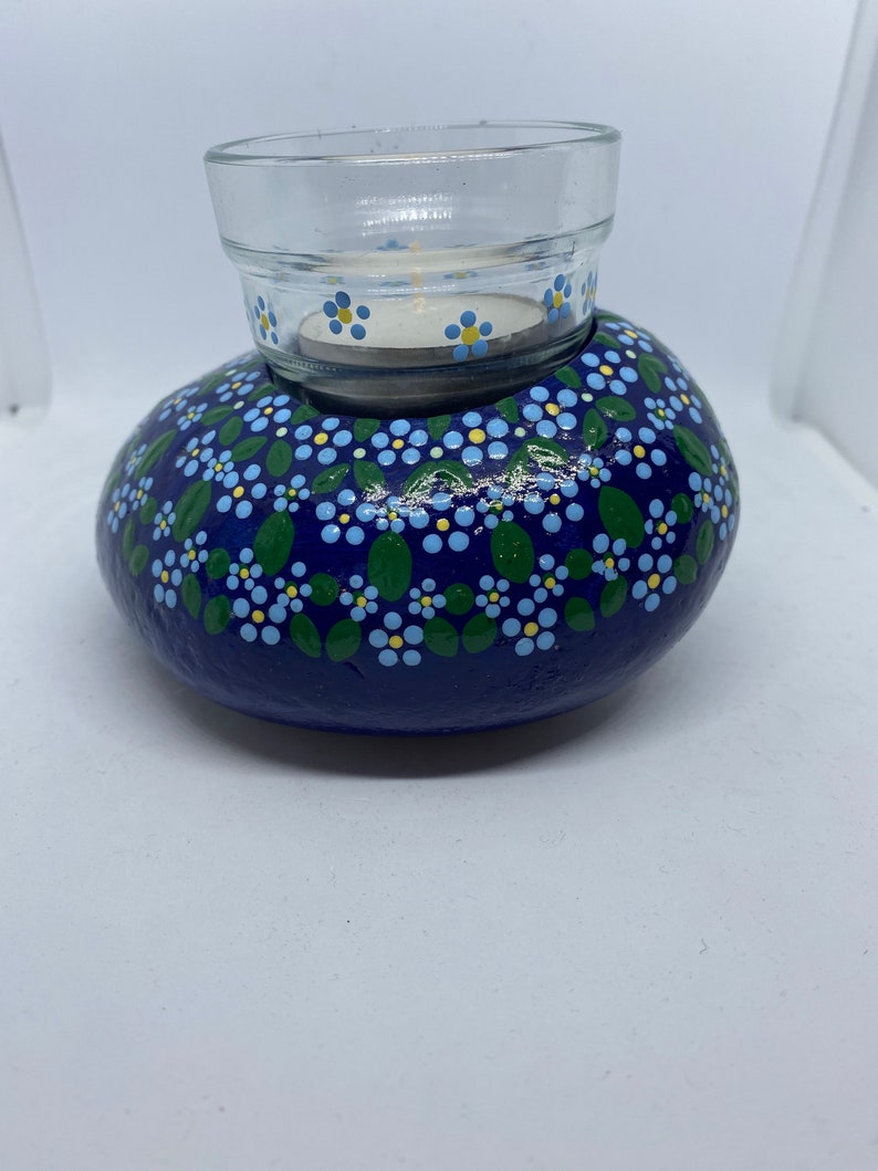 Puede incluir: Una roca pintada de azul con un dise&ntilde;o floral blanco y amarillo, que sostiene un portavelas de vidrio.