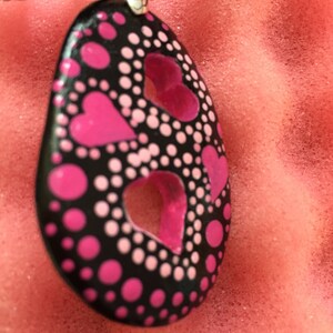 May include: A black pendant necklace with a silver clasp. The pendant is oval-shaped, featuring pink heart cutouts and pink and white polka dots. The necklace cord is black.
