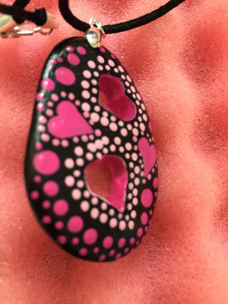 May include: A black pendant necklace with a silver clasp. The pendant is oval-shaped, featuring pink heart cutouts and pink and white polka dots. The necklace cord is black.