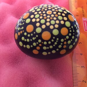 May include: A black stone painted with a mandala design using yellow, orange, and green dots.