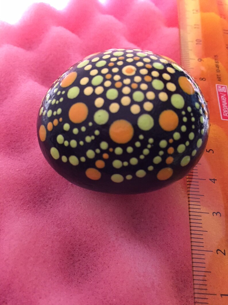 May include: A black stone painted with a mandala design using yellow, orange, and green dots.