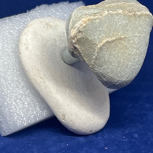 Può includere: Una pietra grigia in equilibrio su una pietra bianca. Le pietre sono lisce e hanno un aspetto naturale e organico.