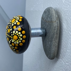 Può includere: Una roccia dipinta di nero e giallo con un motivo a mandala è fissata a una pietra grigia con un gancio di metallo. Questo è un gancio da parete decorativo.