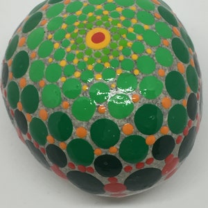 May include: A round, gray stone painted with a mandala design. The mandala features concentric circles of green, yellow, orange, and red dots.