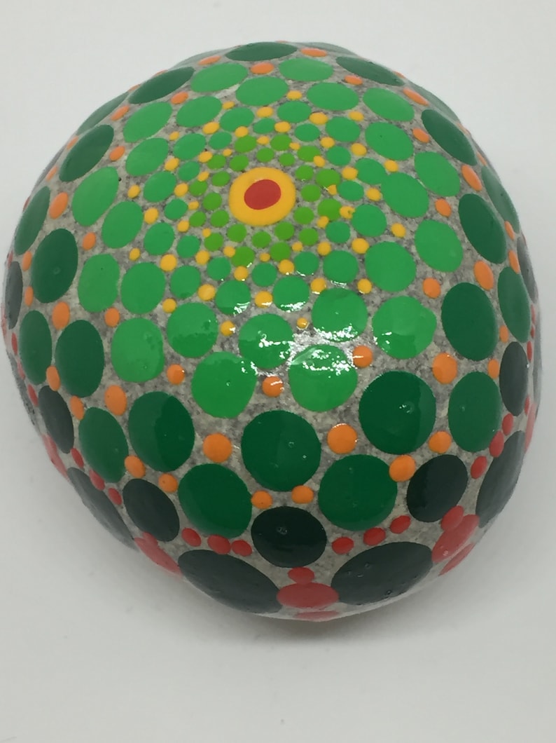 May include: A round, gray stone painted with a mandala design. The mandala features concentric circles of green, yellow, orange, and red dots.