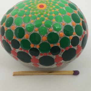 May include: A round, gray stone painted with a mandala design in shades of green, orange, and red. The stone is about the size of a matchstick.