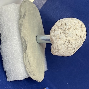 May include: A grey stone with a metal rod attached to a smaller grey stone. The stones are on a white surface.