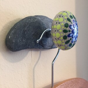 Puede incluir: Un gancho de pared de piedra gris con un pomo redondo pintado. El pomo está decorado con un diseño de mandala en tonos de verde, amarillo, azul y blanco.