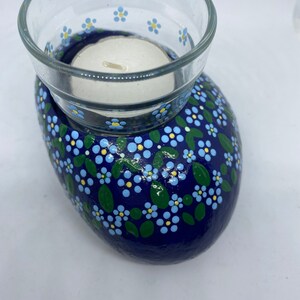 Puede incluir: Una roca pintada de azul con un portavelas para velas de t&eacute; blanco. La roca est&aacute; decorada con flores de nomeolvides azules y blancas y hojas verdes.