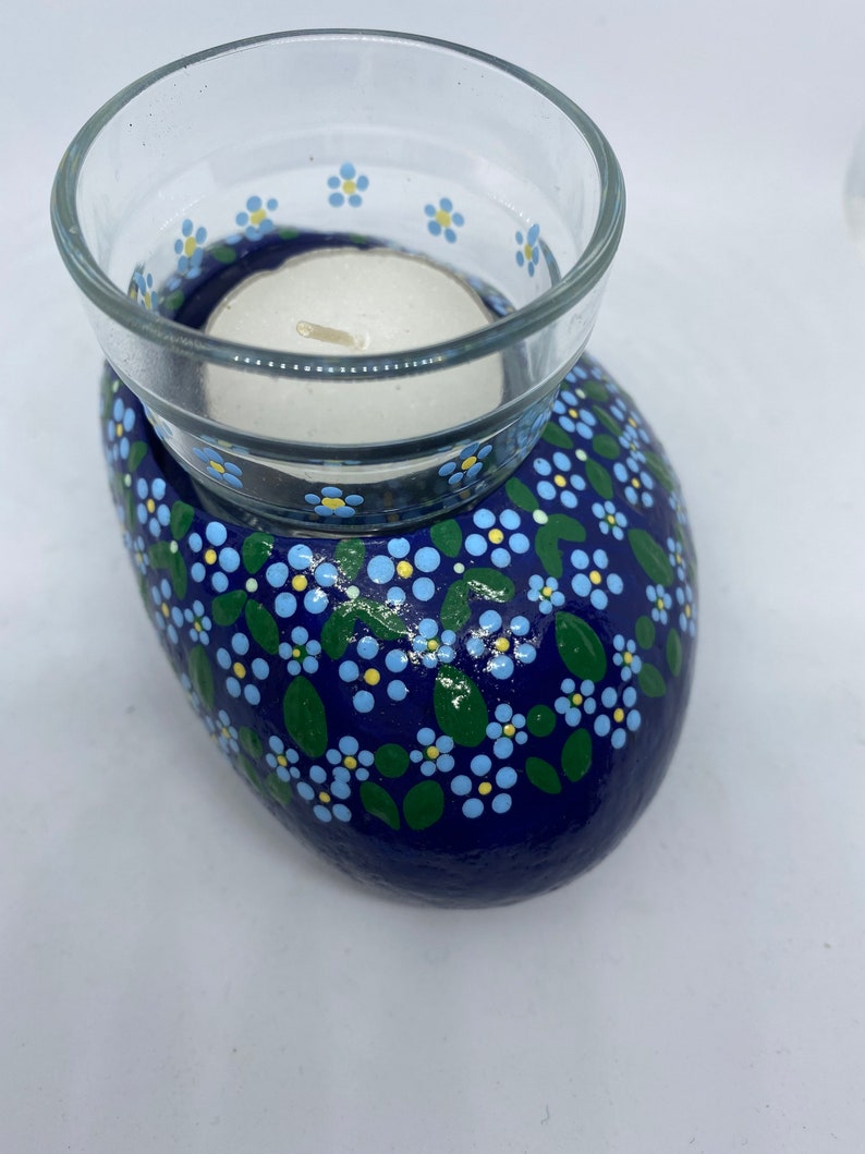 Puede incluir: Una roca pintada de azul con un portavelas para velas de t&eacute; blanco. La roca est&aacute; decorada con flores de nomeolvides azules y blancas y hojas verdes.