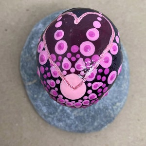 May include: A painted rock with a pink heart design. The rock is covered in small pink and black dots. The heart is outlined in pink and has a pink and white gradient.