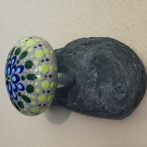 May include: A painted rock mushroom with a green, yellow, blue and white mandala design. The mushroom is attached to a gray rock base.