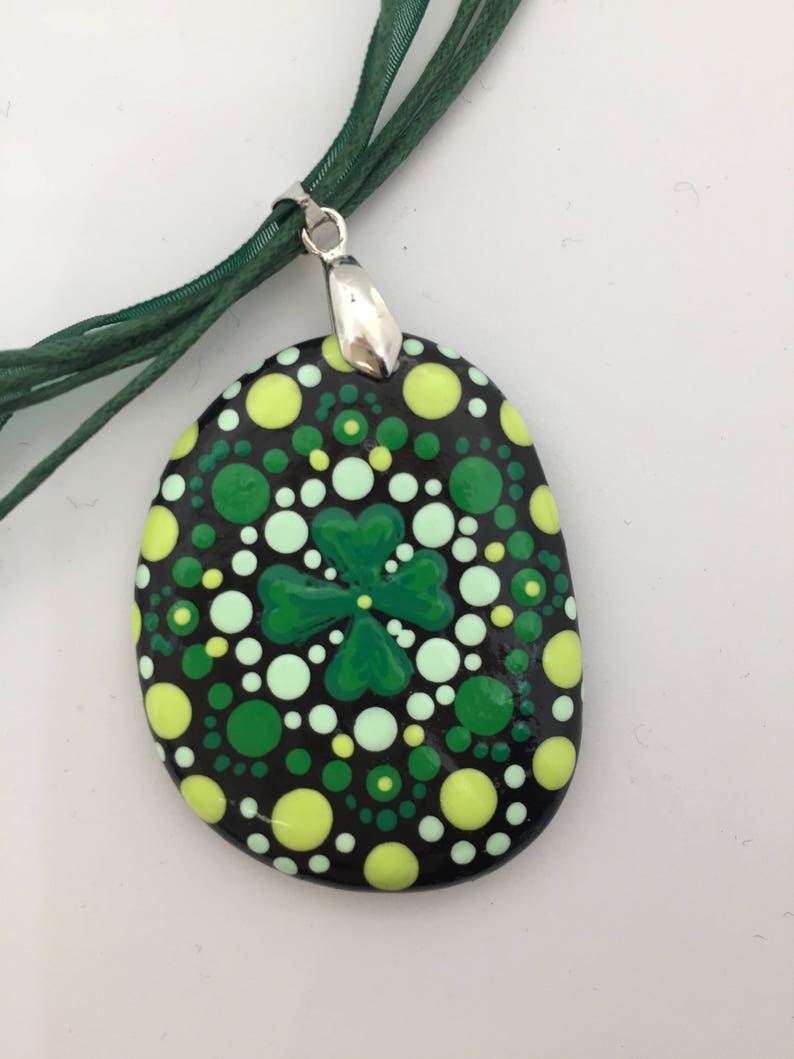 May include: A green four-leaf clover mandala painted on a black stone pendant with a silver bail. The pendant is attached to a green ribbon necklace.