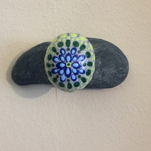 May include: A painted rock with a green, blue, and yellow mandala design. The rock is sitting on top of a larger, gray rock.