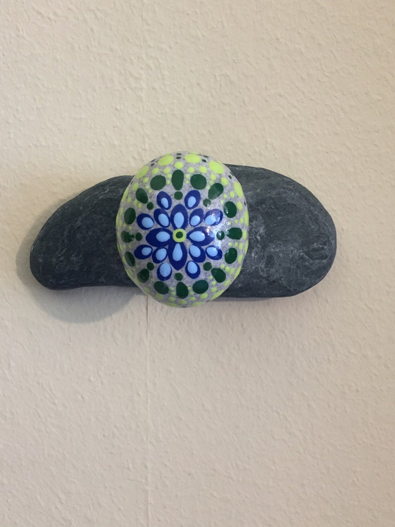 May include: A painted rock with a green, blue, and yellow mandala design. The rock is sitting on top of a larger, gray rock.