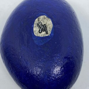 Puede incluir: Una roca pintada de azul con una pegatina blanca. La pegatina tiene las iniciales "SM" escritas con un marcador negro.