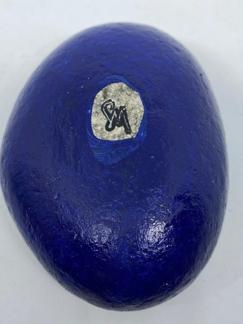 Puede incluir: Una roca pintada de azul con una pegatina blanca. La pegatina tiene las iniciales "SM" escritas con un marcador negro.