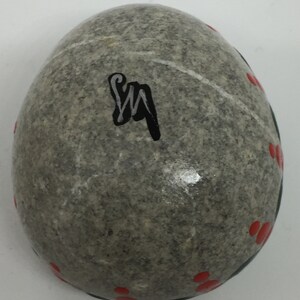 May include: A gray stone egg with red dots and a black signature "SM" painted on the surface.