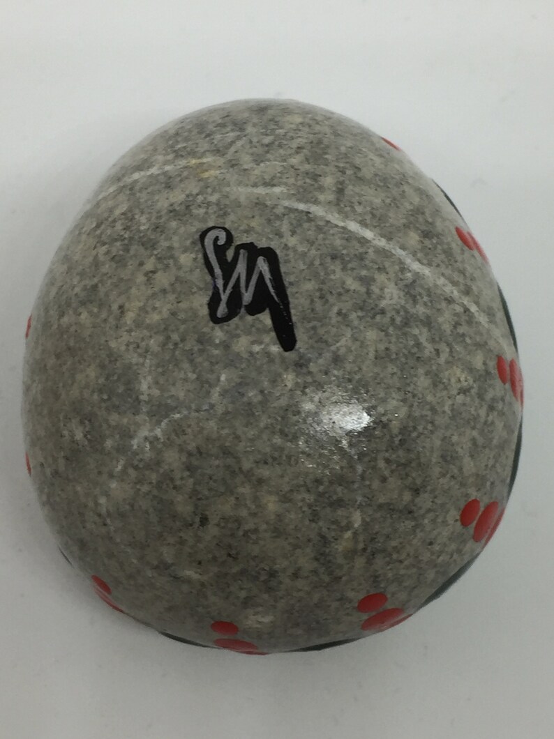 May include: A gray stone egg with red dots and a black signature "SM" painted on the surface.