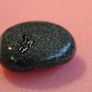 May include: A gray painted rock with white lettering that says "PM 17" and green and red paint accents.