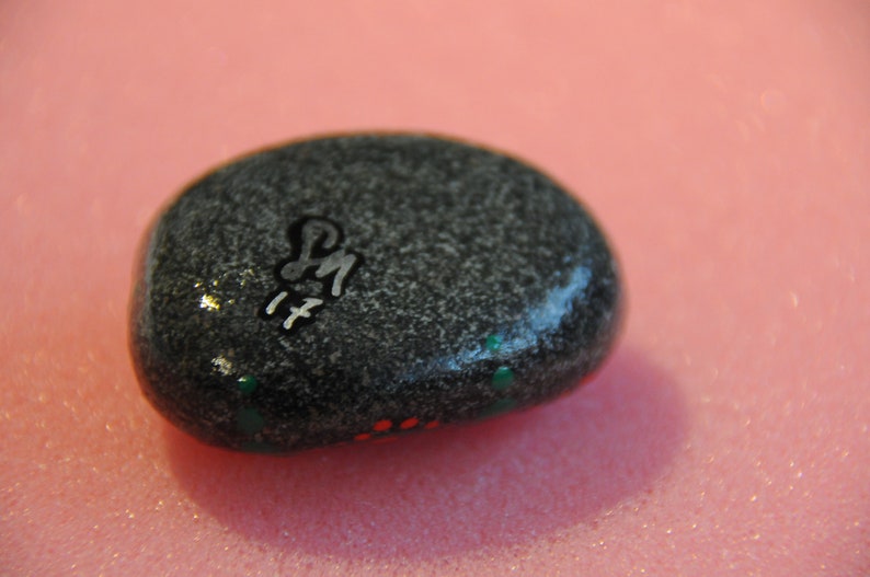 May include: A gray painted rock with white lettering that says "PM 17" and green and red paint accents.