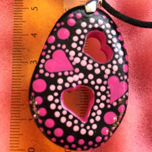 May include: Black pendant necklace with a unique design. The pendant is oval-shaped, featuring pink hearts and polka dots on a black background. The pendant measures approximately 2.5 inches in length and is strung on a black cord.