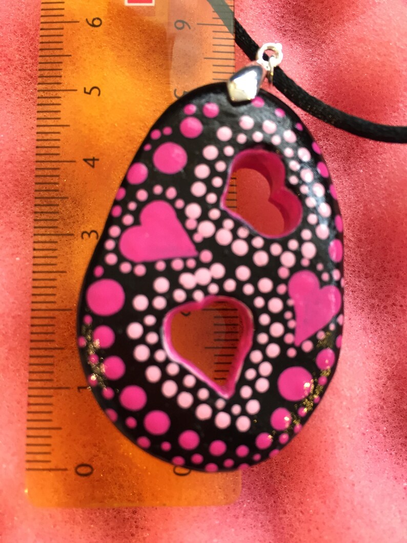 May include: Black pendant necklace with a unique design. The pendant is oval-shaped, featuring pink hearts and polka dots on a black background. The pendant measures approximately 2.5 inches in length and is strung on a black cord.