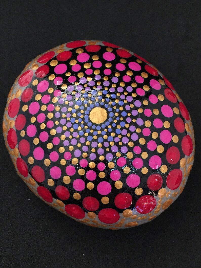 May include: A round, gray stone painted with a mandala design. The mandala features concentric circles of red, pink, purple, and gold dots on a black background.