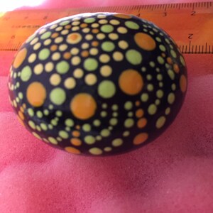 May include: A black stone painted with a mandala design in shades of yellow, orange, and green. The stone is round and smooth.