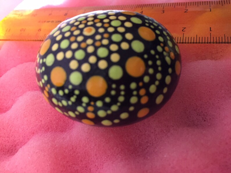 May include: A black stone painted with a mandala design in shades of yellow, orange, and green. The stone is round and smooth.