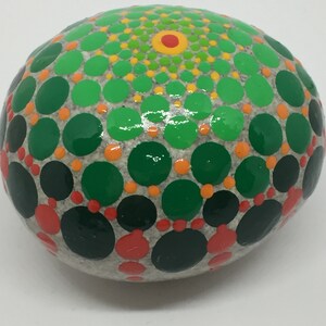 May include: A round, gray stone painted with a green, orange, and red mandala design. The mandala is made up of concentric circles of dots.