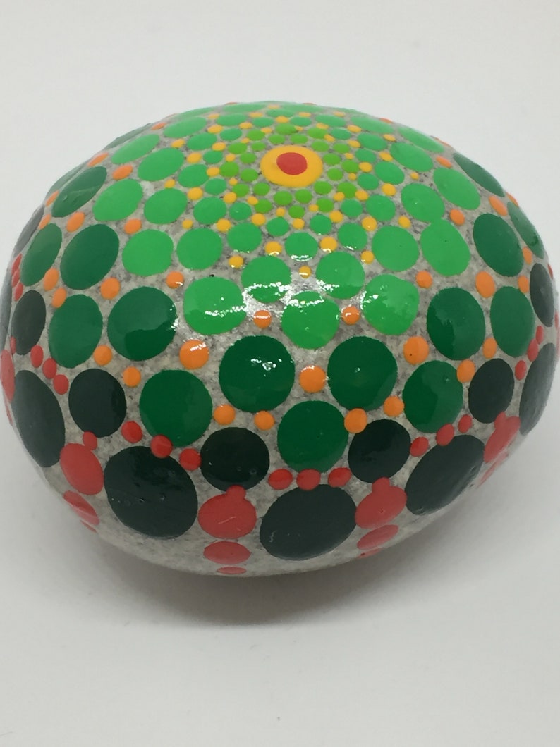 May include: A round, gray stone painted with a green, orange, and red mandala design. The mandala is made up of concentric circles of dots.