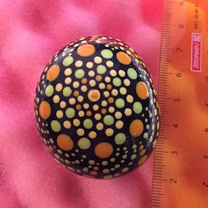 May include: A black stone painted with a mandala design in shades of orange, yellow, and green. The stone is about 2 inches in diameter.