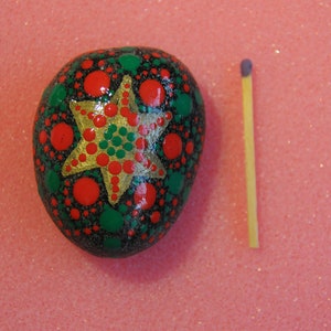 May include: A hand-painted rock with a green, red, and gold star design. The rock is about the size of a matchstick.