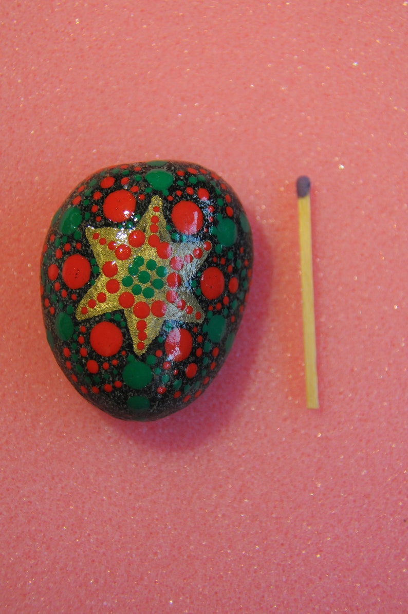 May include: A hand-painted rock with a green, red, and gold star design. The rock is about the size of a matchstick.