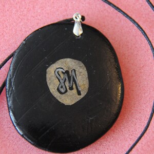 May include: Black pendant necklace with a silver bail and a gold-colored circle with the initials "SM" in silver lettering.