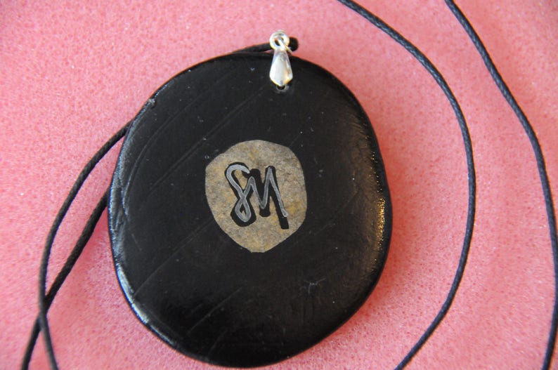May include: Black pendant necklace with a silver bail and a gold-colored circle with the initials "SM" in silver lettering.