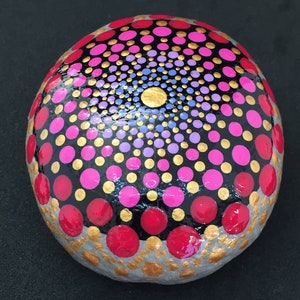 May include: A round, gray stone painted with a mandala design. The mandala features concentric circles of pink, purple, gold, and red dots on a black background.
