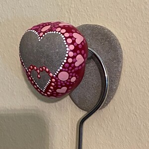 May include: A grey stone painted with pink and red dots and hearts, attached to a silver hook. The stone is mounted on a grey stone base.
