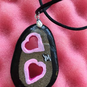 May include: A hand-painted stone pendant necklace. The black, oval-shaped stone features two pink hearts with white outlines. The pendant hangs from a black cord with a silver-colored clasp. A small silver-colored initial "M" is also on the stone.