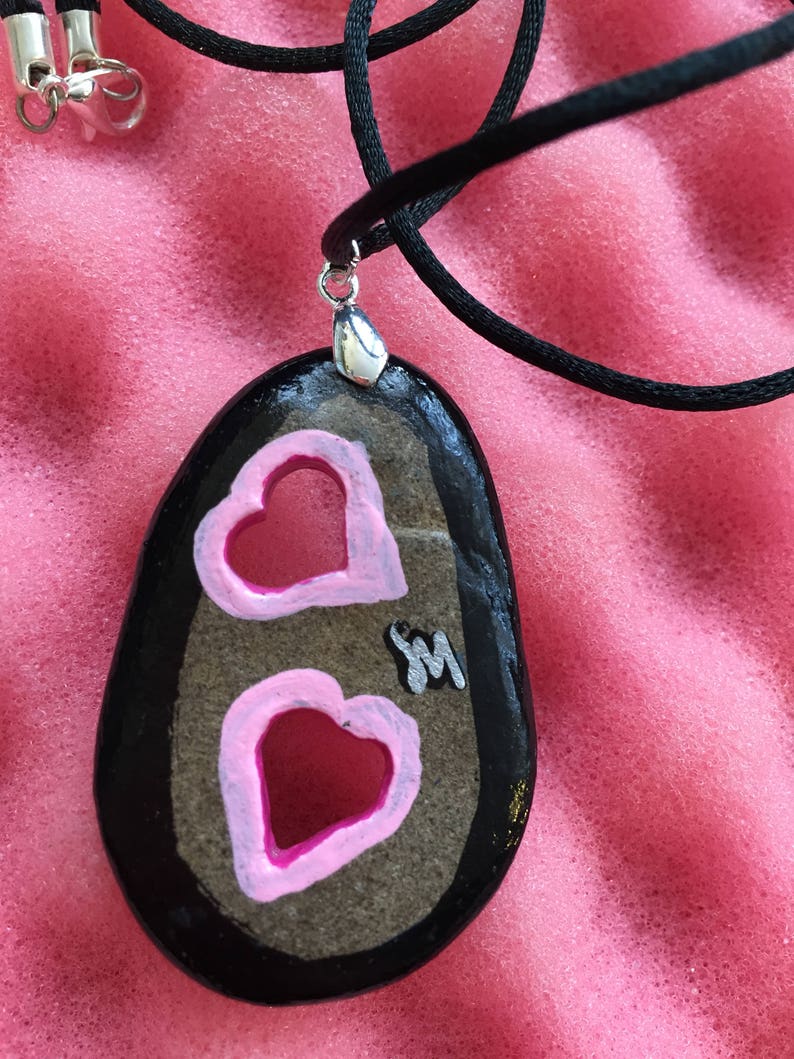 May include: A hand-painted stone pendant necklace. The black, oval-shaped stone features two pink hearts with white outlines. The pendant hangs from a black cord with a silver-colored clasp. A small silver-colored initial "M" is also on the stone.