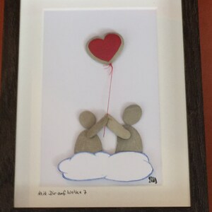 May include: A framed pebble art piece depicting two figures holding a red heart-shaped balloon. The figures are standing on a white cloud with a blue outline. The text "Mit Dir auf Wolke 7" is written below the cloud.