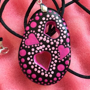 May include: Black pendant with pink hearts and polka dots, strung on a black cord. The pendant has two heart-shaped cutouts and a silver-colored bail. The design features a mix of large and small pink dots.