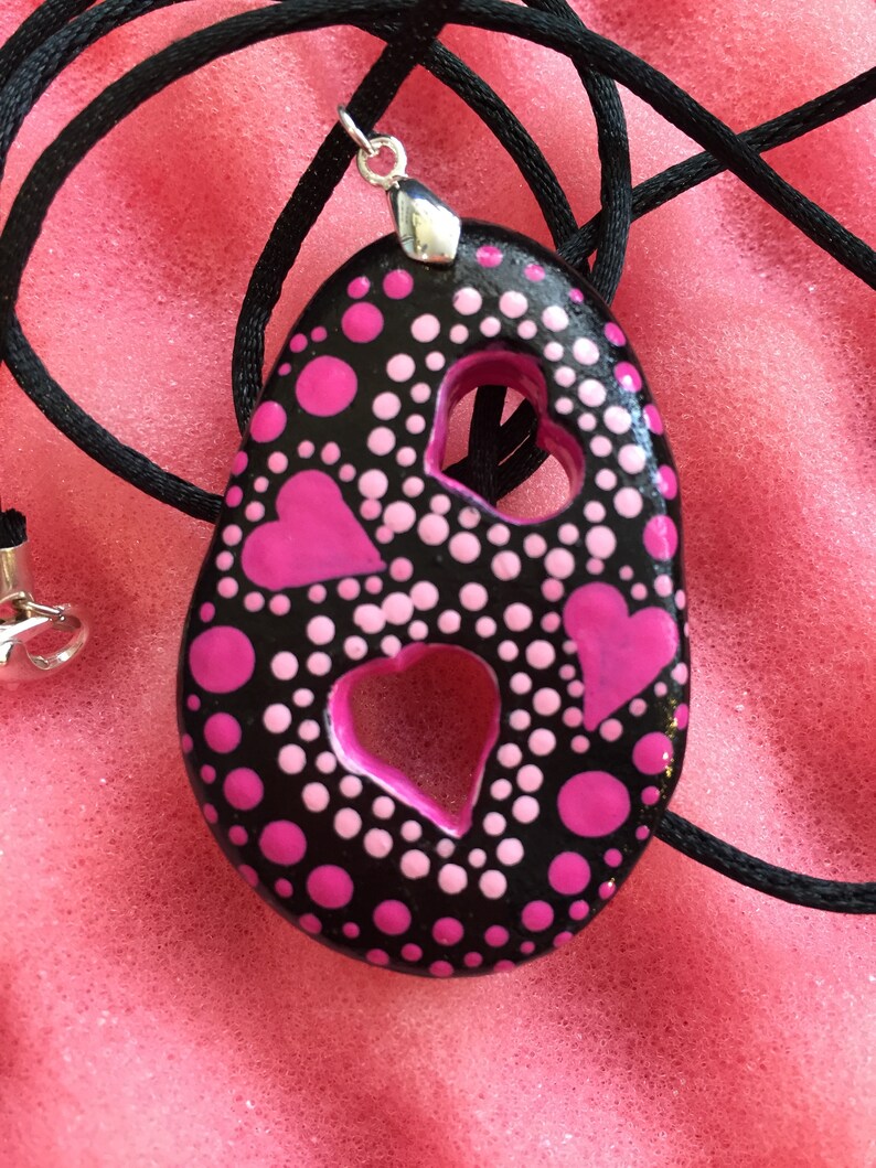 May include: Black pendant with pink hearts and polka dots, strung on a black cord. The pendant has two heart-shaped cutouts and a silver-colored bail. The design features a mix of large and small pink dots.