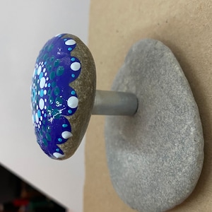 Op de afbeelding: Een beschilderde stenen paddenstoel met een blauw, wit en turquoise mandala-ontwerp. De paddenstoel is bevestigd aan een grijze stenen basis met een metalen staaf.