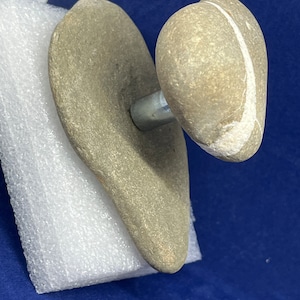 Può includere: Una maniglia per porta unica realizzata con pietre naturali. La maniglia presenta un pomello in pietra arrotondato con una striscia bianca e una base piatta a forma di cuore. Le pietre sono di colore grigio chiaro e la maniglia è fissata con un'asta di metallo argentato.