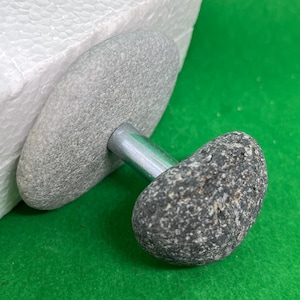 Può includere: Un pomo in pietra grigia con una barra di metallo argentata attaccata a una base in pietra grigia. Il pomo è attaccato a una superficie bianca.