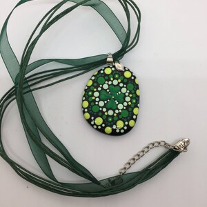 May include: A green and yellow painted rock pendant with a four-leaf clover design, hanging from a green ribbon necklace.